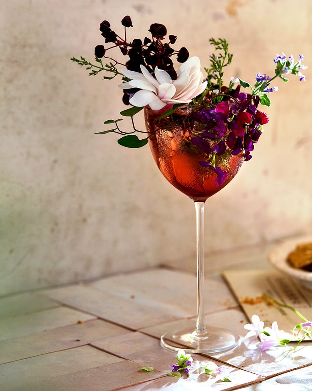 Close-up of a stemmed wine glass filled with red liquid and decorated with various flowers, set against a light, textured background.