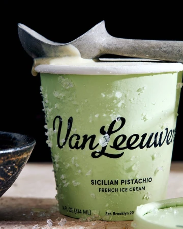 Close-up shot of Van Leeuwen's Sicilian Pistachio French Ice Cream in a green paper container with a metal ice cream scoop.