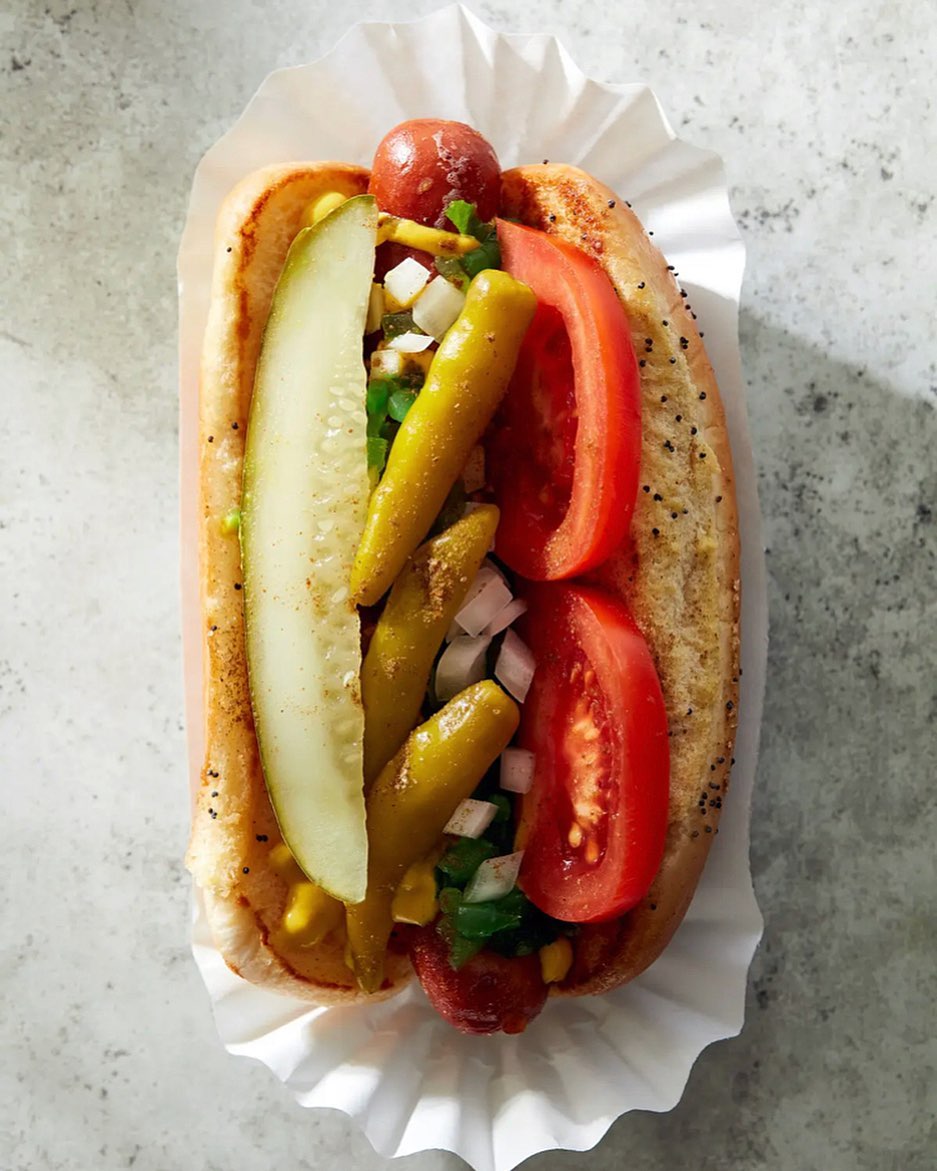 Overhead shot of a loaded hot dog with sliced tomatoes, pickles, and peppers in a bun, served on white paper on a grey surface.
