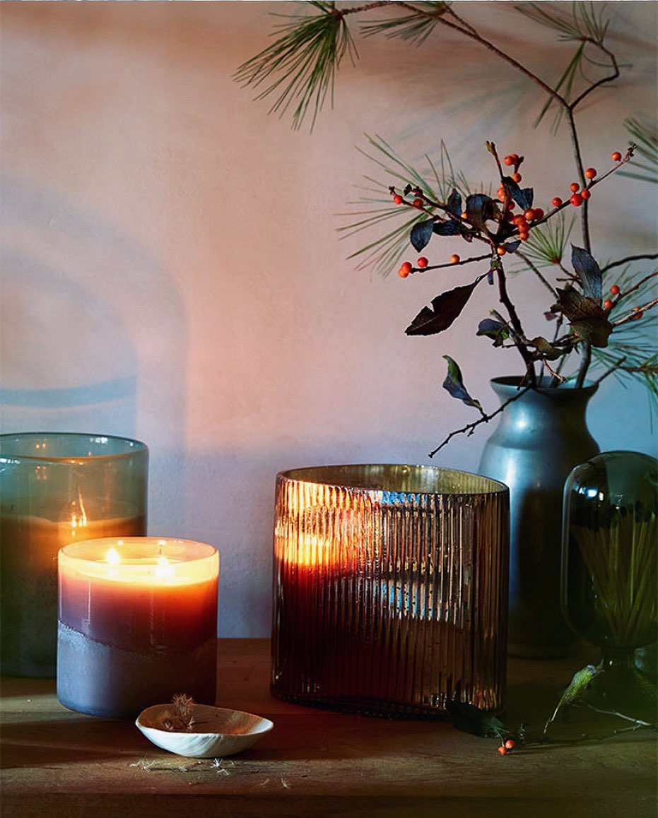 Candlelight illuminates a room with two glass-encased candles and a vase of winter berries, offering a warm, inviting ambiance.