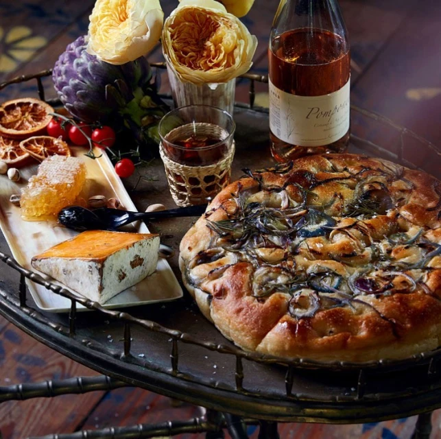 Overhead close-up still life of focaccia with onion and herbs, cheese, honeycomb, and a bottle and glass of drink on a metal tray.