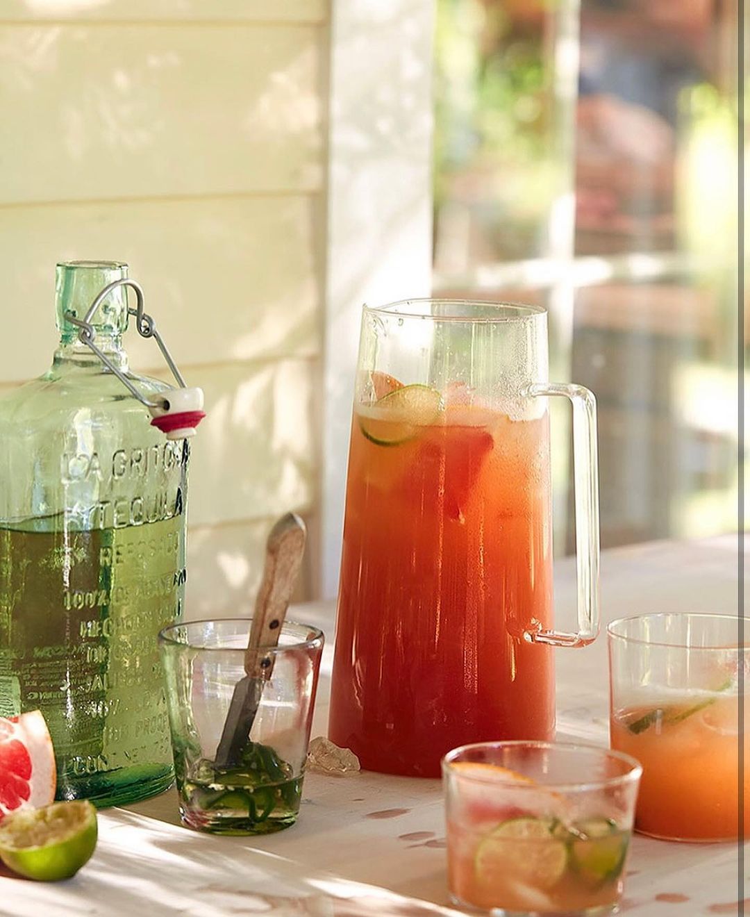 Clear glass pitcher of grapefruit cocktail, with other glassware and fruit slices, on a white table in bright daylight.