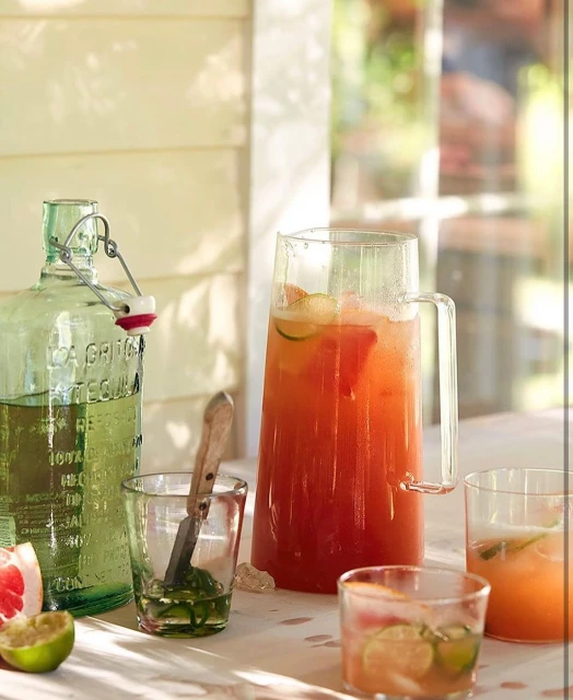 Clear glass pitcher of grapefruit cocktail, with other glassware and fruit slices, on a white table in bright daylight.