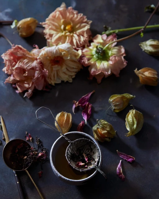 Still life photography of dahlia flowers, golden berries, tea strainer, and cup with tea on dark surface. Moody, artistic composition.