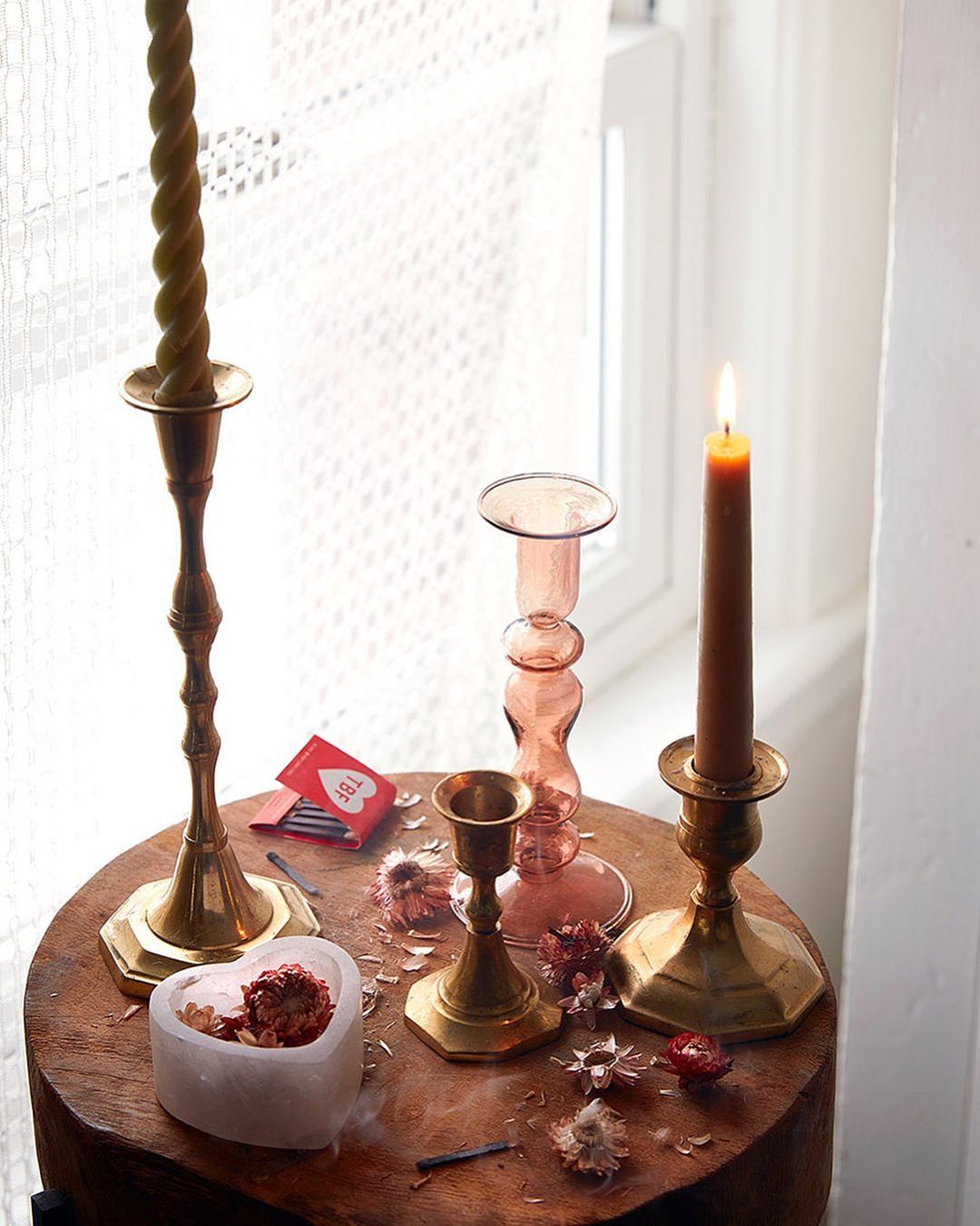 Several lit and unlit candles sit on wood surface near window. Candles are in brass and glass candle holders. Dried flowers scattered around.