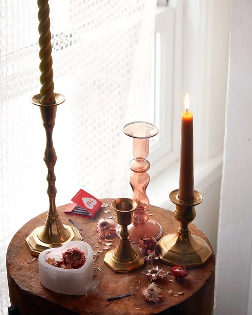Several lit and unlit candles sit on wood surface near window. Candles are in brass and glass candle holders. Dried flowers scattered around.
