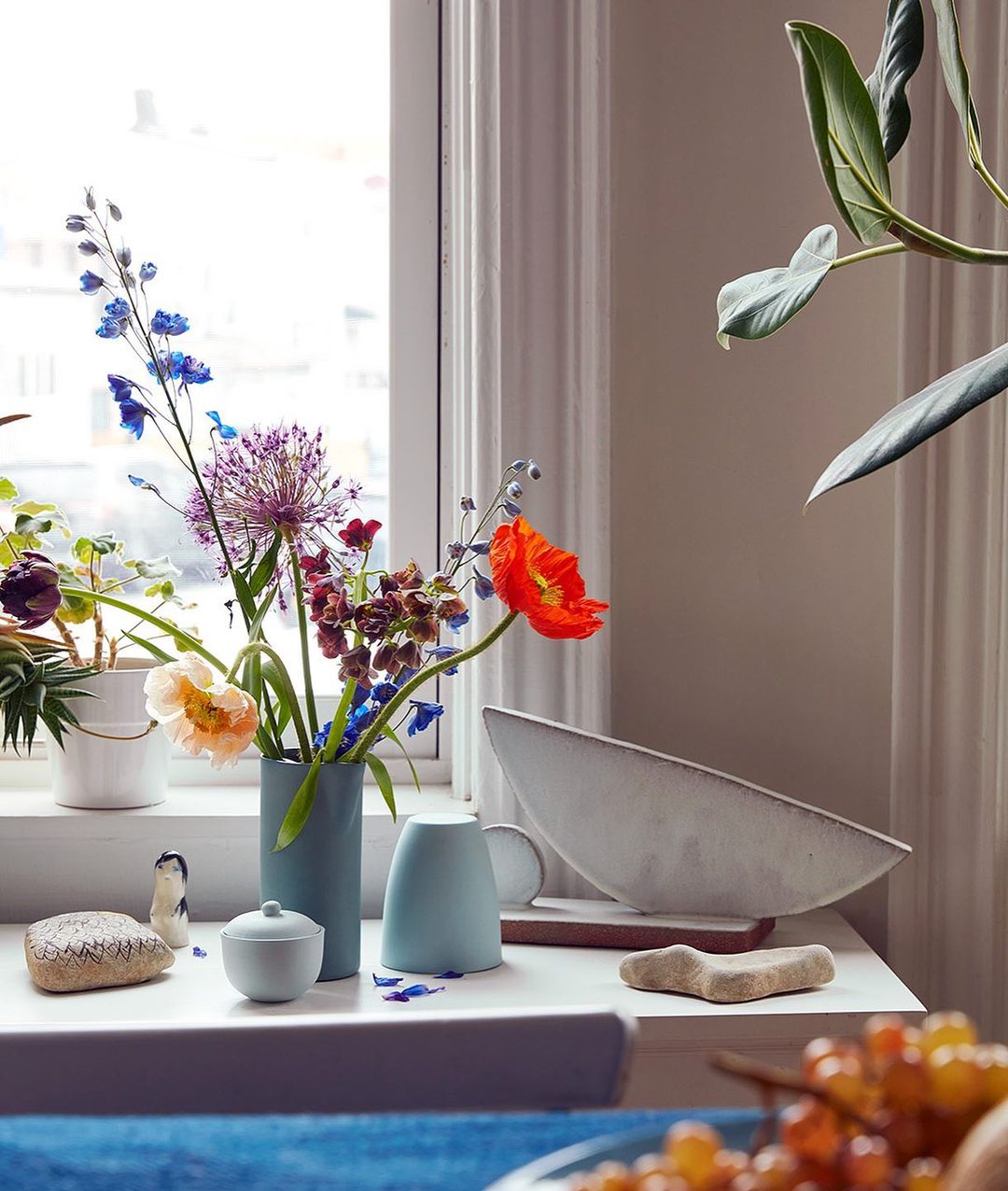 Still life on a white table featuring a blue vase with colorful flowers next to a decorative bowl and other small objects, indoors.