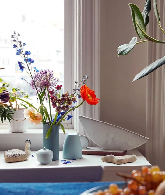 Still life on a white table featuring a blue vase with colorful flowers next to a decorative bowl and other small objects, indoors.