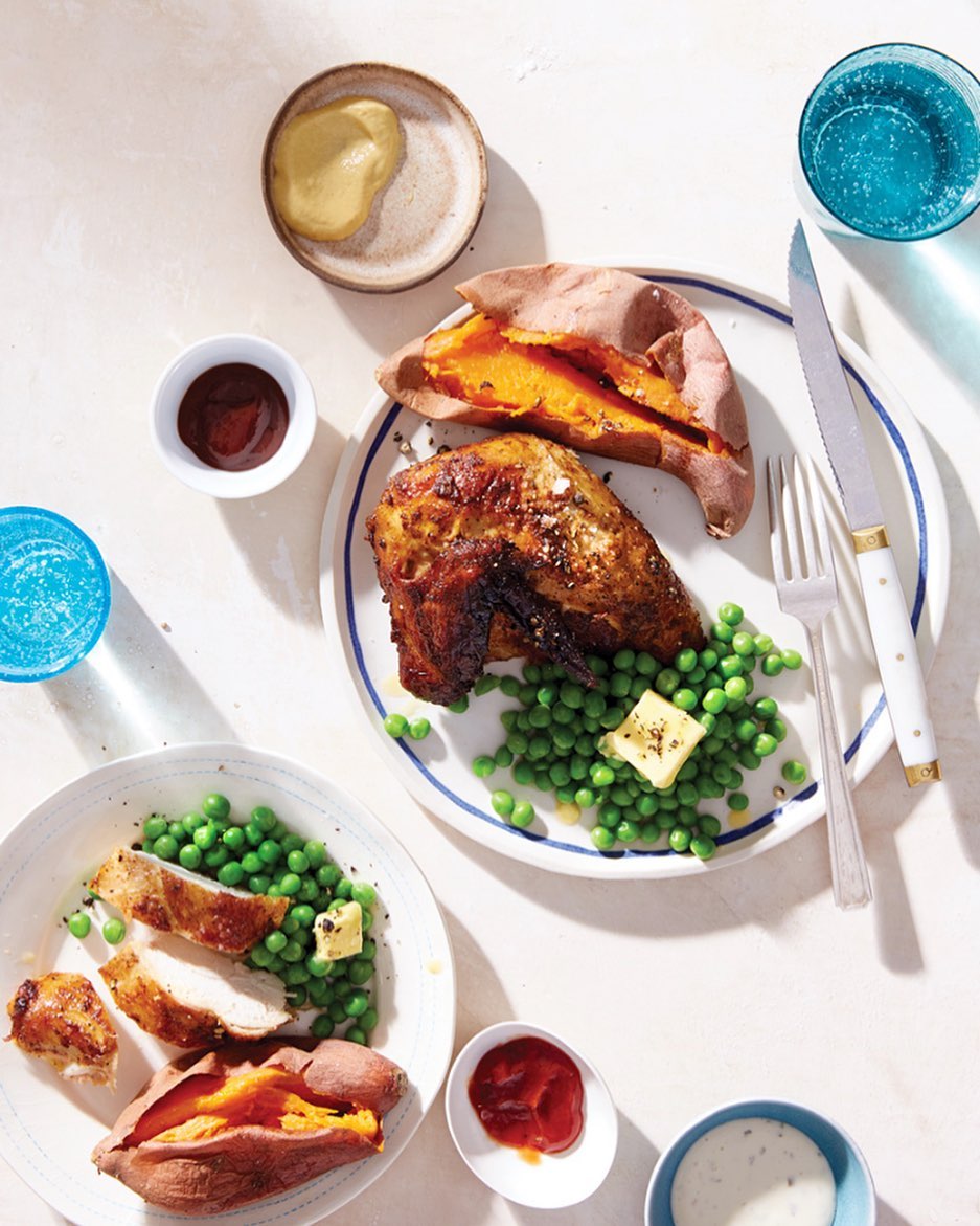 Overhead shot of plates of chicken and sweet potatoes with sides of peas and dipping sauces on a white table, bright, natural light.