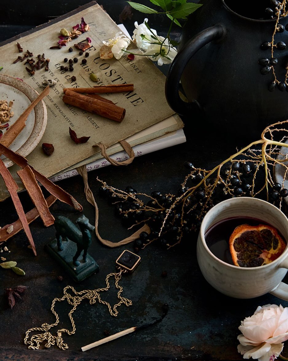 Still life featuring a mug of mulled wine with an orange slice, spices, vintage books, jewelry, a bronze horse, and flower blooms.