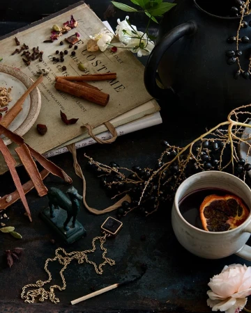 Still life featuring a mug of mulled wine with an orange slice, spices, vintage books, jewelry, a bronze horse, and flower blooms.