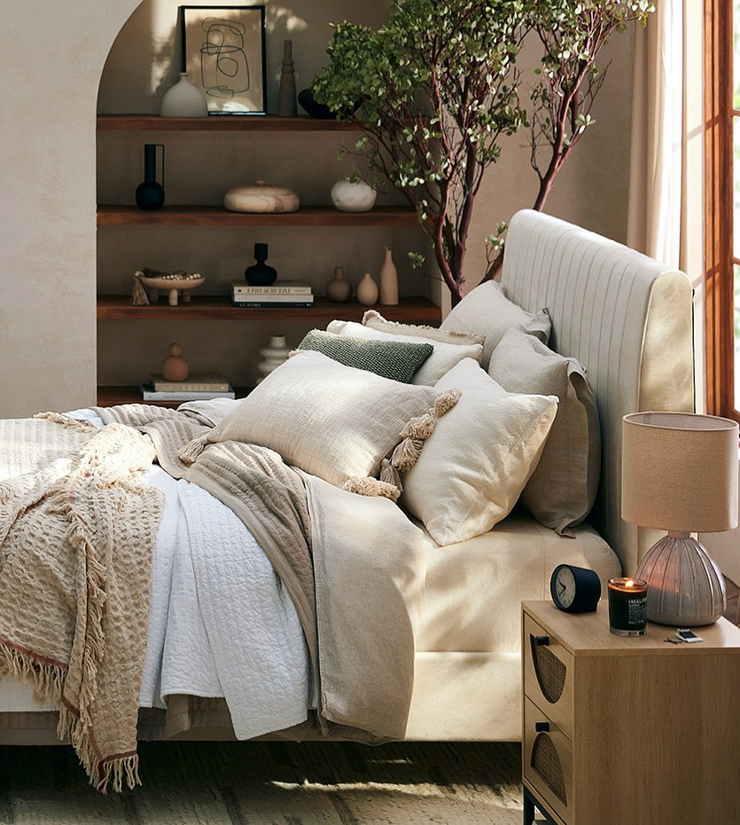 Bedroom interior with neutral tones featuring a bed with many pillows, a nightstand, lamp, and decorative objects in a calm setting.
