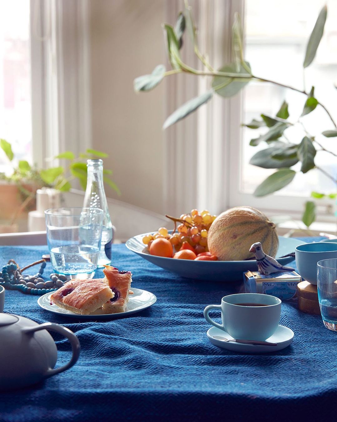 Still life featuring a blue table cloth with cake, fruit, drinks, and a teacup placed in a natural light setting near a window.