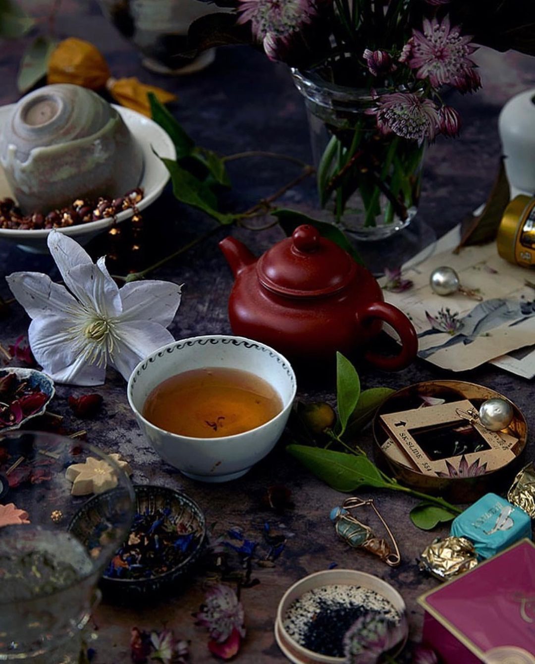 Tea set with dark brown teapot, cup of tea, and purple flowers in glass vase, surrounded by scattered chocolates and rose petals.