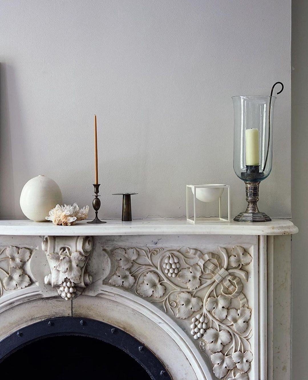 Ornate mantelpiece with decorative carvings and antique objects, including candles, crystal, and a glass hurricane lamp, in a home interior.