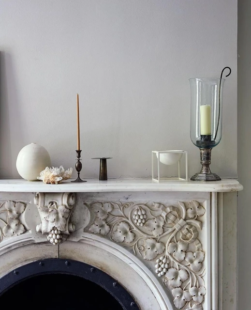 Ornate mantelpiece with decorative carvings and antique objects, including candles, crystal, and a glass hurricane lamp, in a home interior.