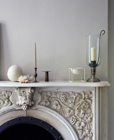 Ornate mantelpiece with decorative carvings and antique objects, including candles, crystal, and a glass hurricane lamp, in a home interior.