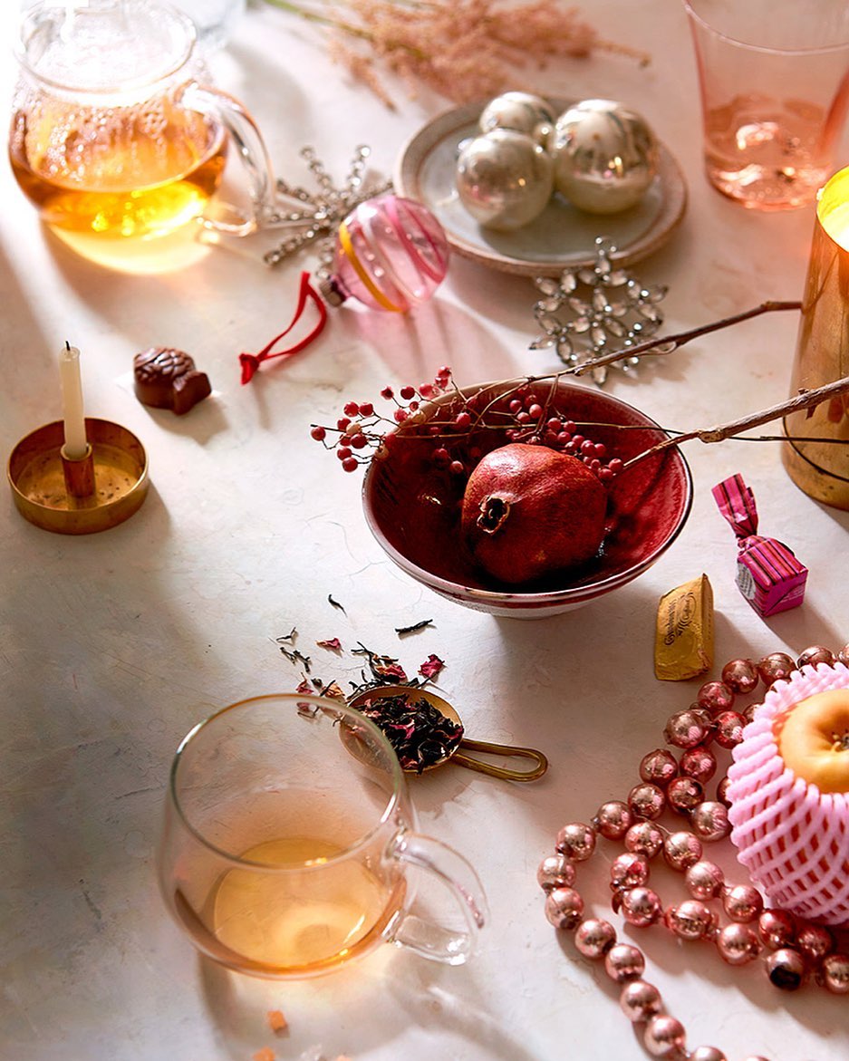 Festive still life featuring a glass teapot, ornaments, a pomegranate in a red bowl, chocolates, and string of beads on a white surface.