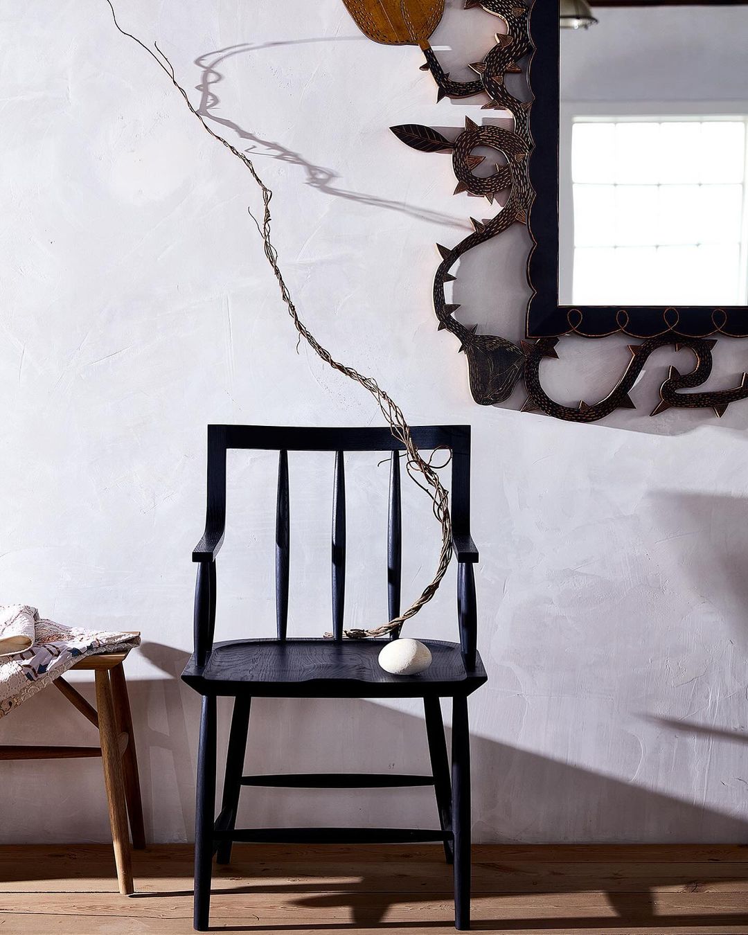 Black wooden chair with a white rock, set against a stark white wall with a decorative mirror, creating a minimalist, artistic composition.