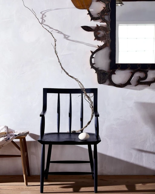 Black wooden chair with a white rock, set against a stark white wall with a decorative mirror, creating a minimalist, artistic composition.