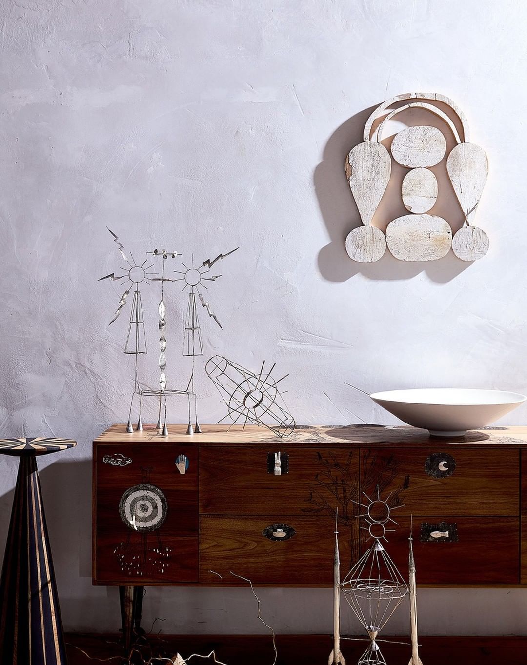 A collection of abstract metal sculptures displayed on a wooden cabinet against a textured light grey wall, with a white bowl on top.