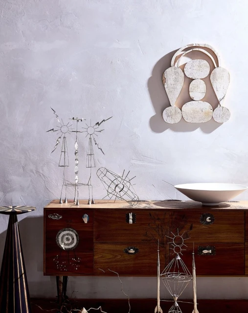 A collection of abstract metal sculptures displayed on a wooden cabinet against a textured light grey wall, with a white bowl on top.