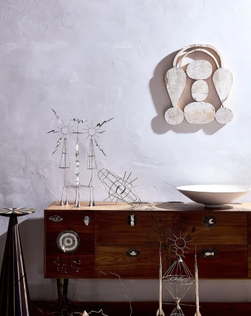 A collection of abstract metal sculptures displayed on a wooden cabinet against a textured light grey wall, with a white bowl on top.
