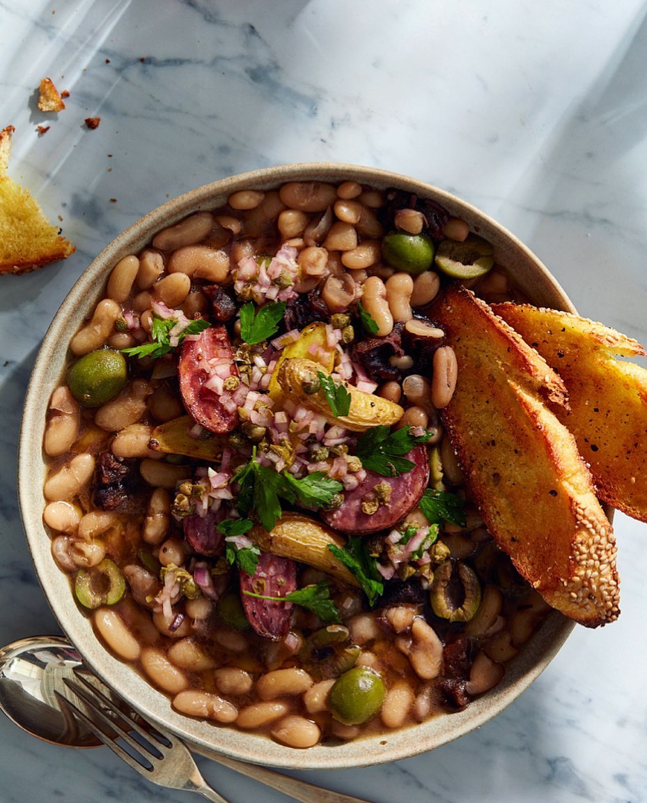 Overhead shot of a bowl of white beans with slices of sausage, green olives, and toasted bread alongside it on a marble surface.