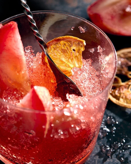 A close-up studio shot of a clear glass filled with crushed ice, topped with slices of fruit and a drink stirrer.