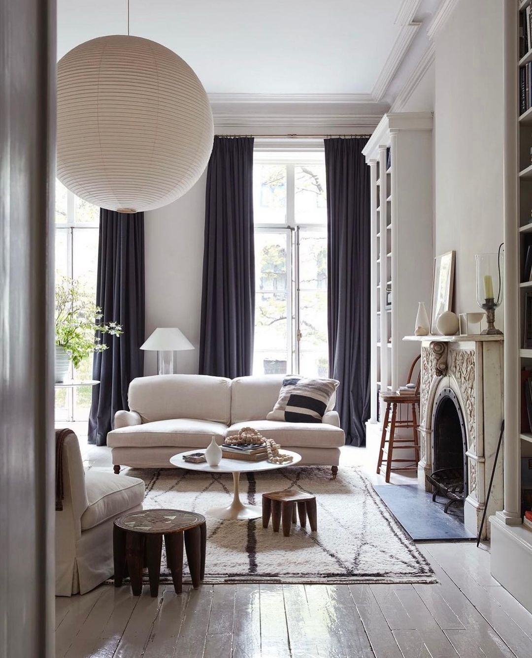 Living room interior with white sofa, round coffee table, wooden stools, area rug, fireplace, white walls, bookshelves and natural light.
