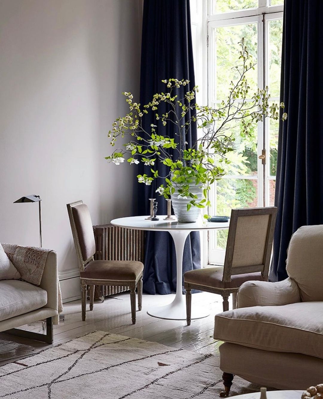 A bright indoor living space features two tan upholstered armchairs, a white round tulip table with chairs, and neutral walls, lit by daylight.