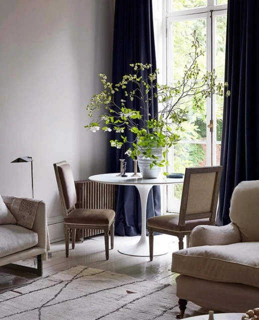A bright indoor living space features two tan upholstered armchairs, a white round tulip table with chairs, and neutral walls, lit by daylight.