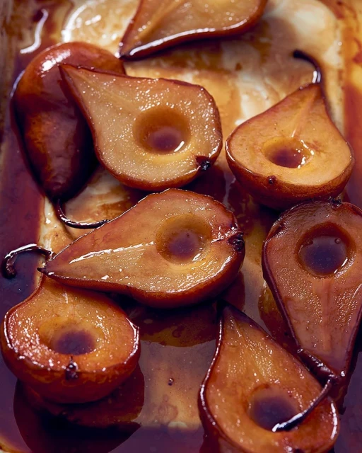 Close-up of halved poached pears in syrup, arranged in a baking dish. The pears are golden-brown and glistening, ready to serve.