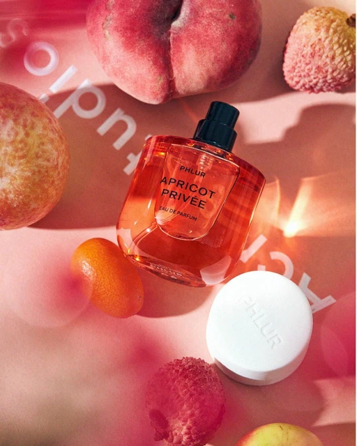 Phlur Apricot Privee perfume bottle surrounded by peaches, lychees, and kumquats on a soft pink surface. Warm, inviting fragrance.