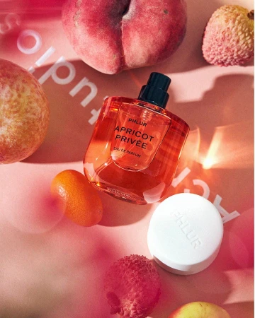 Phlur Apricot Privee perfume bottle surrounded by peaches, lychees, and kumquats on a soft pink surface. Warm, inviting fragrance.