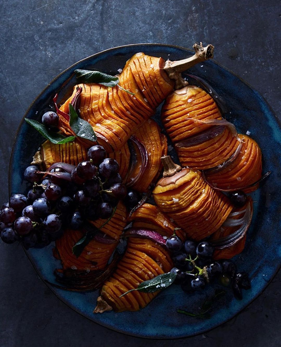 Close-up of roasted butternut squash with dark grapes, onions, and herbs on a rustic blue plate against a dark grey backdrop.