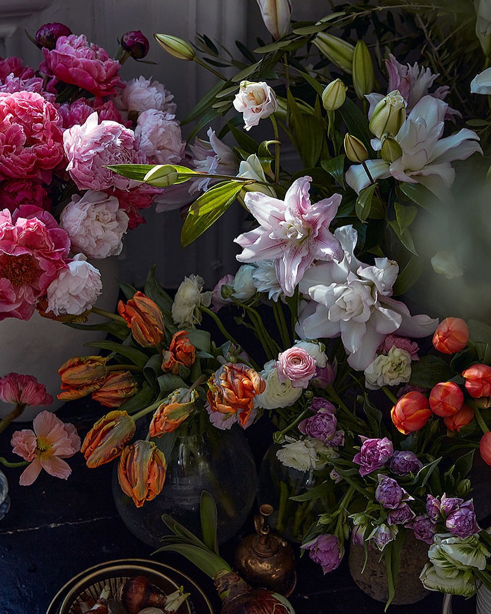 Floral arrangement featuring pink peonies, lilies, and tulips in glass vases, displayed on a dark table against a neutral background.