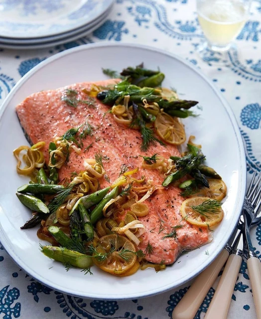 Overhead shot of salmon fillet on white plate, garnished with asparagus, dill, and lemon slices, displayed on blue patterned tablecloth.
