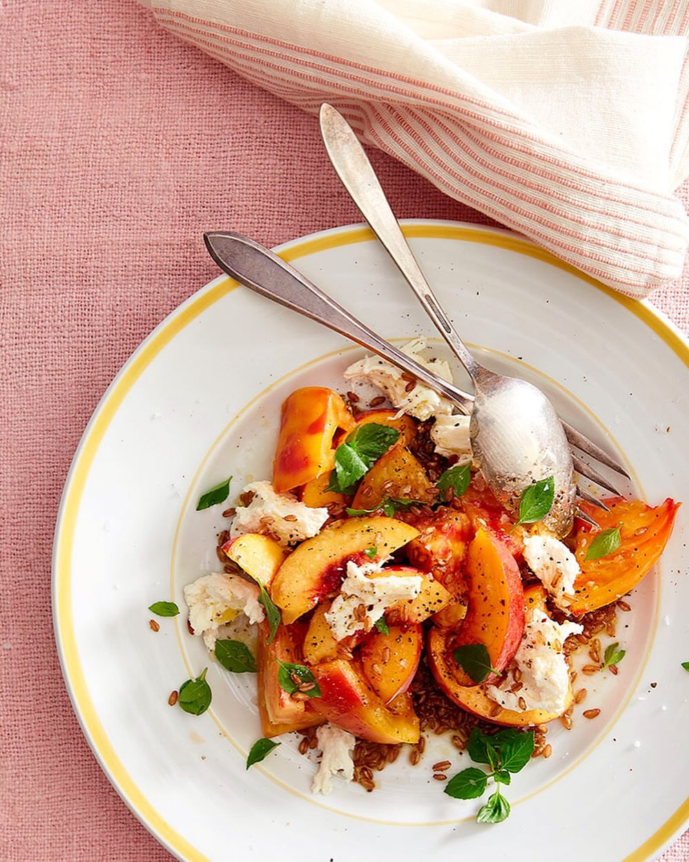 Overhead shot of a white plate with a peach and tomato salad, featuring fresh basil and cheese, served on a pink striped cloth.