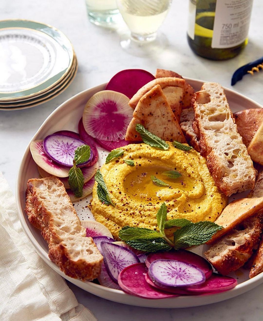 Overhead shot showcasing a bowl of hummus surrounded by sliced radishes, pita bread, and herbs, accompanied by wine and plates.