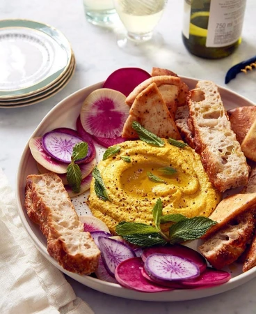 Overhead shot showcasing a bowl of hummus surrounded by sliced radishes, pita bread, and herbs, accompanied by wine and plates.