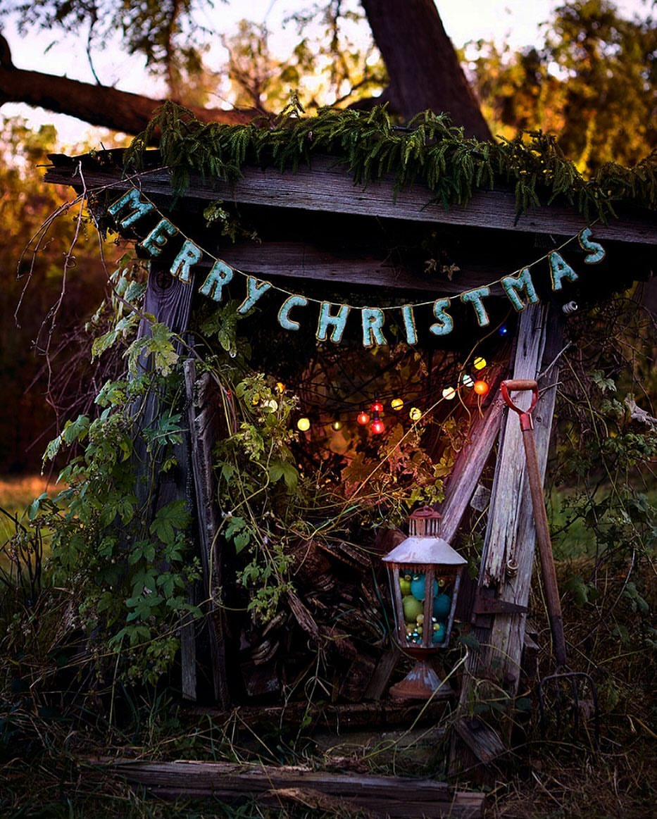 Outdoor Christmas display featuring a weathered wooden structure adorned with a 'Merry Christmas' banner, string lights, and lush greenery.