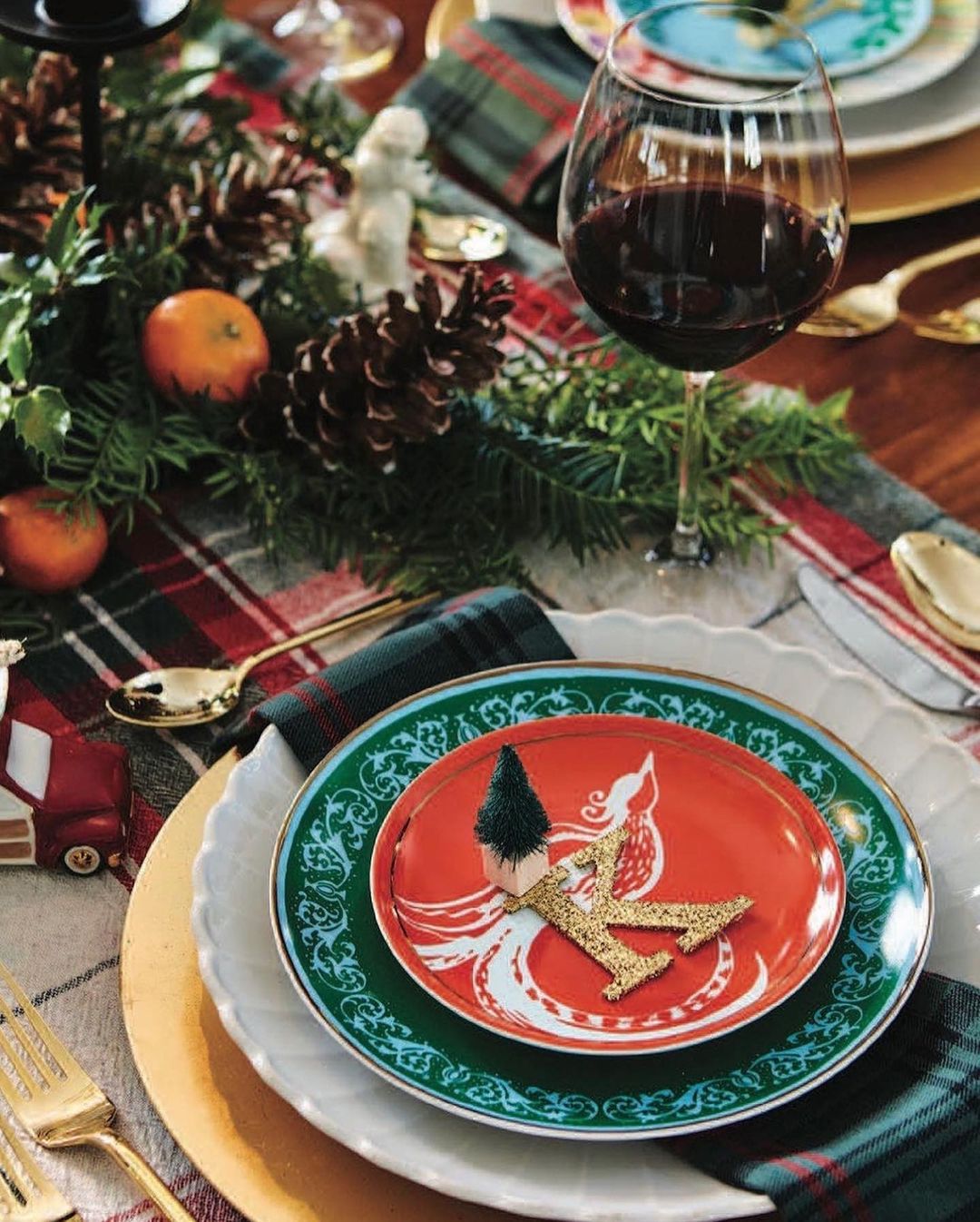 Festive table setting with red wine, ornaments, pinecones, candles, tableware, and tartan runner for a holiday meal.