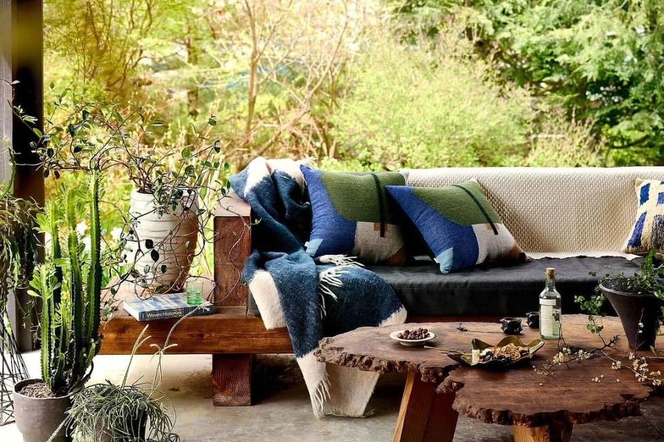 Outdoor living space featuring a wooden sofa with patterned cushions and a throw blanket, complemented by unique wooden tables and potted plants.