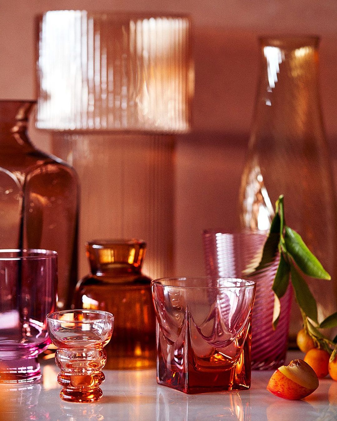Still life shot of an assortment of colourful glass vases, bottles, and glasses in pink, orange, and brown tones against a pink background.