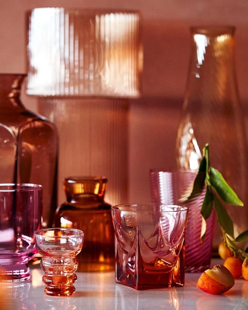 Still life shot of an assortment of colourful glass vases, bottles, and glasses in pink, orange, and brown tones against a pink background.