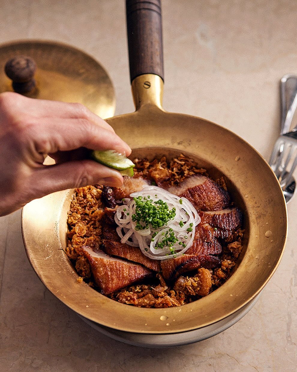 Overhead shot of char siu pork in a gold pan, topped with sliced onion and greens, being drizzled with lime juice.