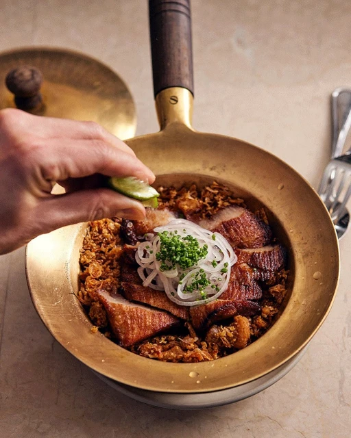 Overhead shot of char siu pork in a gold pan, topped with sliced onion and greens, being drizzled with lime juice.