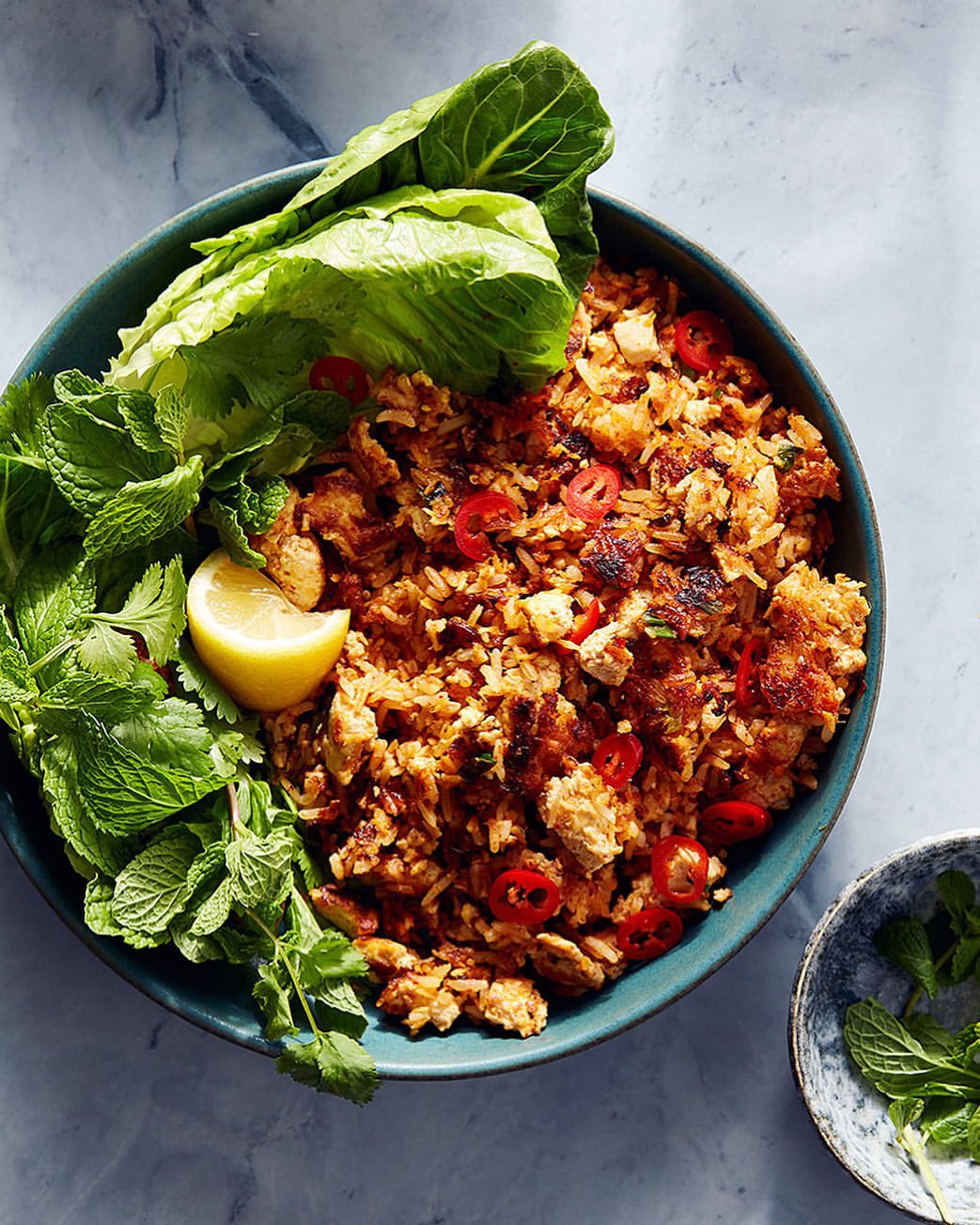 Overhead shot of appetizing fried rice dish in teal bowl, garnished with fresh mint, lettuce, lemon, red chili pepper slices.