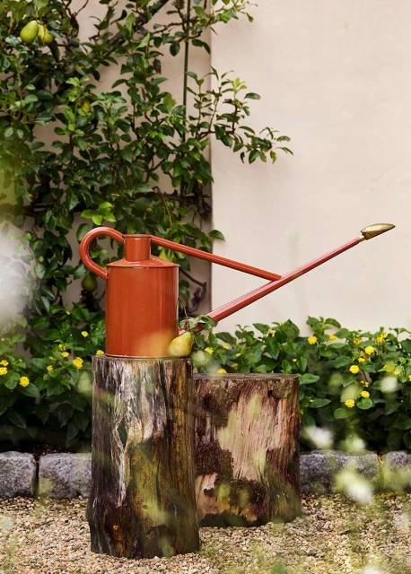 Orange watering can on wooden tree stump in garden setting with foliage and wall. Outdoors in natural light. Rustic garden decor.